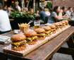 Ten cheeseburgers with lettuce, tomato, pickles, onions, and melted cheese on sesame seed buns on a long wooden board